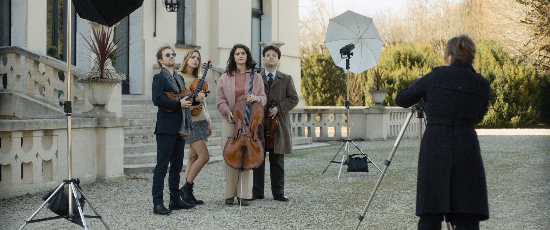 Four musicians posing with a cello and violins outdoors while a photographer shoots them.