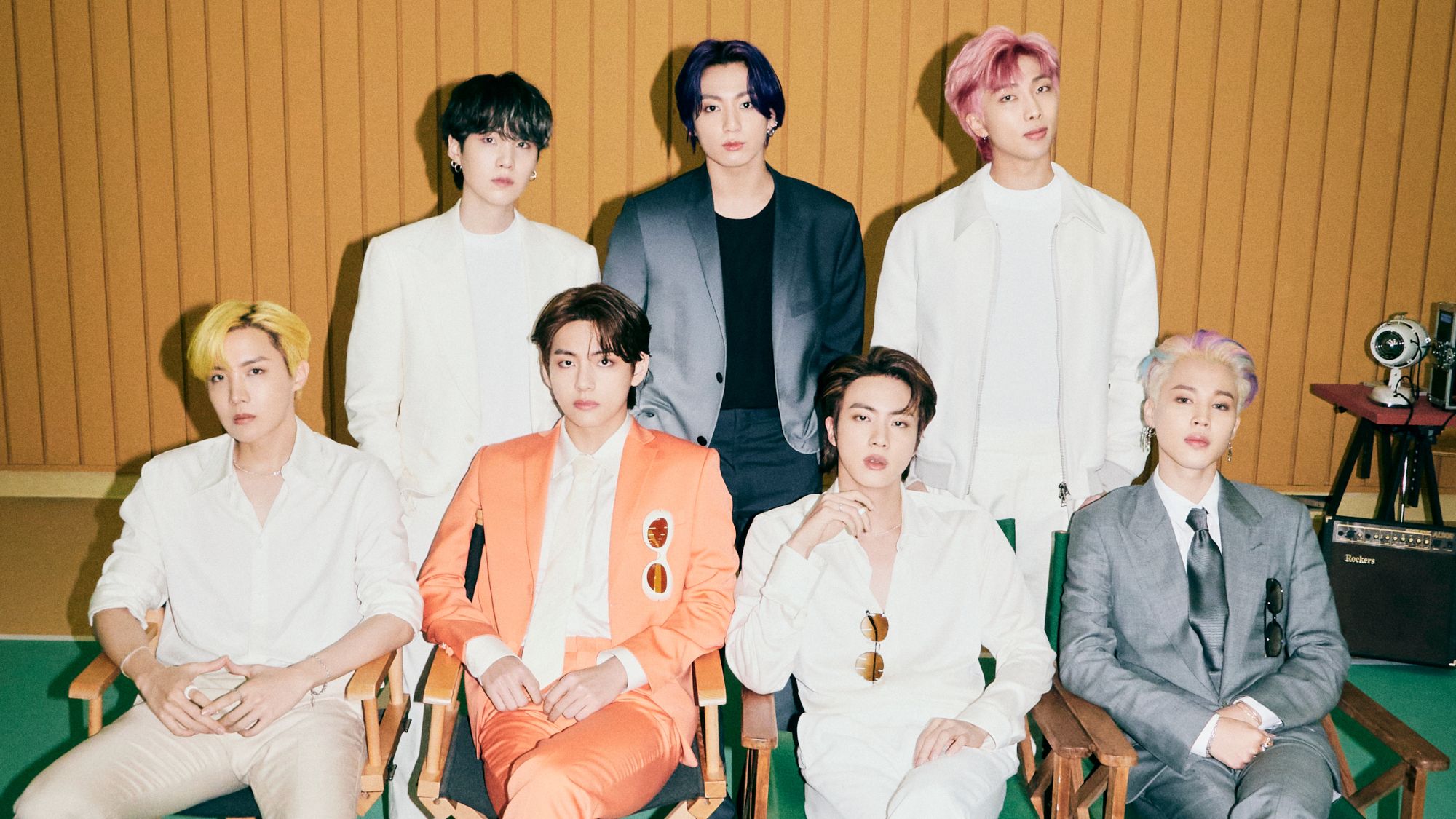 entertainment-musik-bts-aufm1 - IN München - Das Stadtmagazin Seven men in colorful suits seated and standing in a studio with a wooden paneled wall, posing for a group photo.
