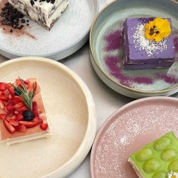 fluffyfuwa.pancake_2 - IN München - Das Stadtmagazin Assorted small desserts on pastel plates: a purple square cake with a yellow flower, a green rectangular cake with round toppings, a strawberry-topped beige pastry, a pink plate with a white dessert, and a white dessert with red dusting.