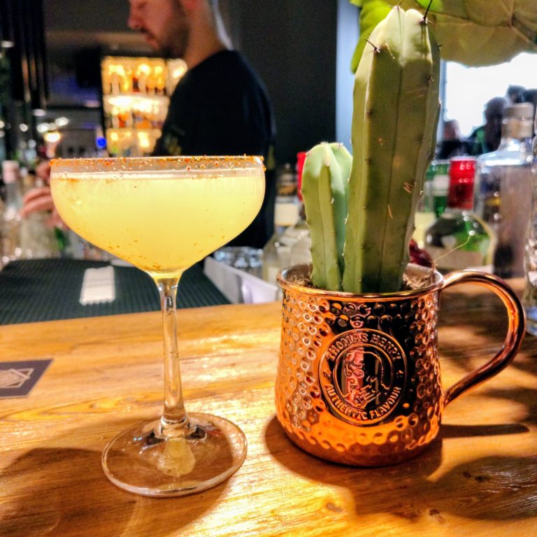 frida1-aw-768x768 - IN München - Das Stadtmagazin Pale yellow cocktail in a coupe glass on a wooden bar next to a hammered copper mug with a green cactus.