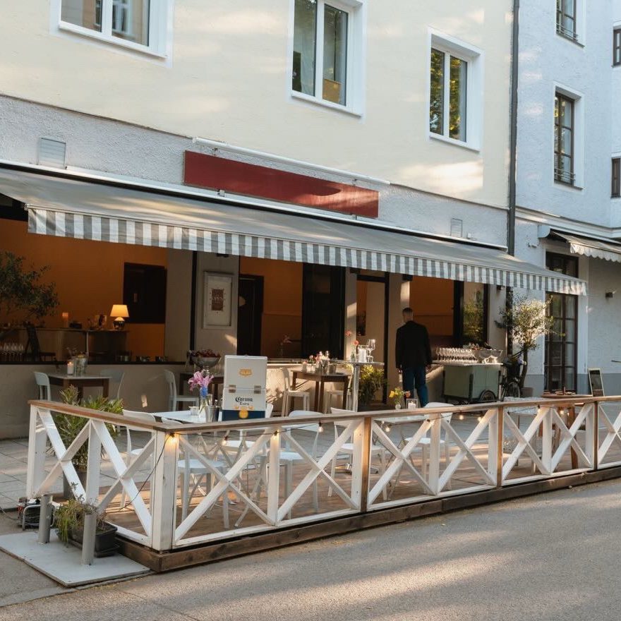 iamfloriphotography und ninety5studios (4) - IN München - Das Stadtmagazin Outdoor restaurant terrace with white crisscross railing, striped awning, and tables set for dining along a sidewalk.