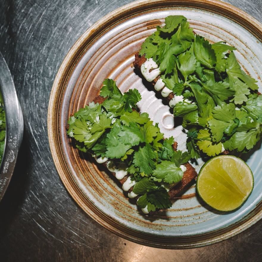 iamfloriphotography und ninety5studios - IN München - Das Stadtmagazin Two cilantro-topped tacos with a lime wedge on a blue-rimmed plate, viewed from above on a metal counter.