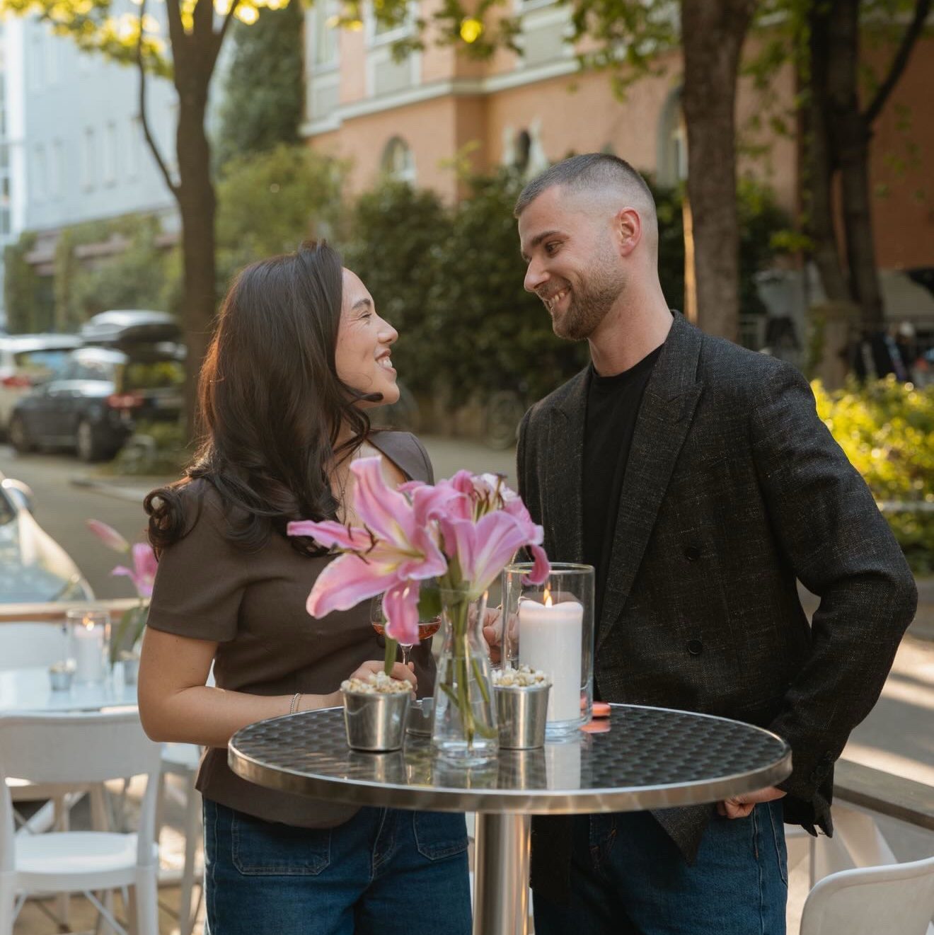 ninety5studios - IN München - Das Stadtmagazin Smiling couple at an outdoor cafe standing near a round metal table with pink lilies, candles, and drinks, talking warmly to each other.