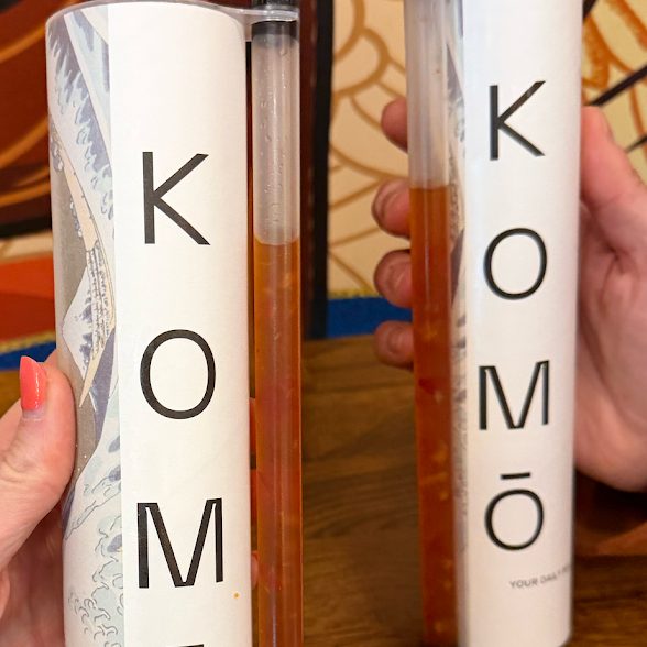 sonia raeder - IN München - Das Stadtmagazin Two tall white boxes labeled KOMÖ on a wooden table, each with a transparent glass tube containing orange liquid held by hands.