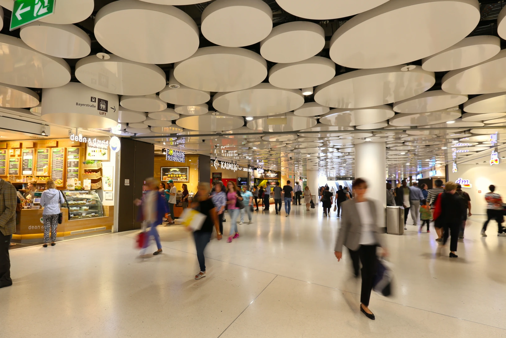 Busy indoor shopping concourse with circular white ceiling panels and people walking past food stalls on the left.