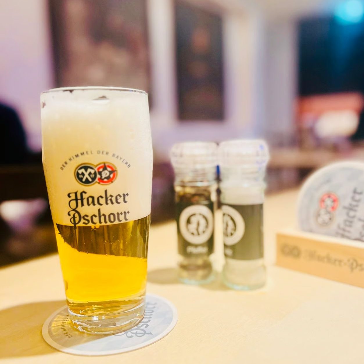 w=1253 - IN München - Das Stadtmagazin Tall glass of pale Hacker-Pschorr beer with a foamy head on a bar counter; logo visible on the glass.