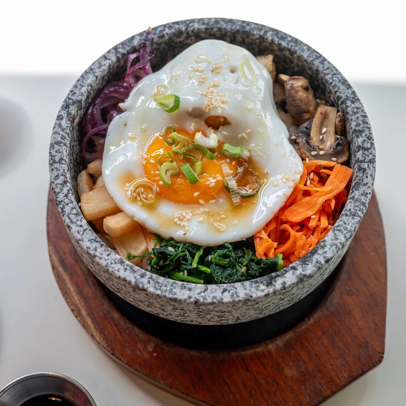 w=1367 (1) - IN München - Das Stadtmagazin Stone bowl bibimbap with a fried egg on top, sesame, scallions, and assorted veggies like carrots, mushrooms, and greens on a wooden board.