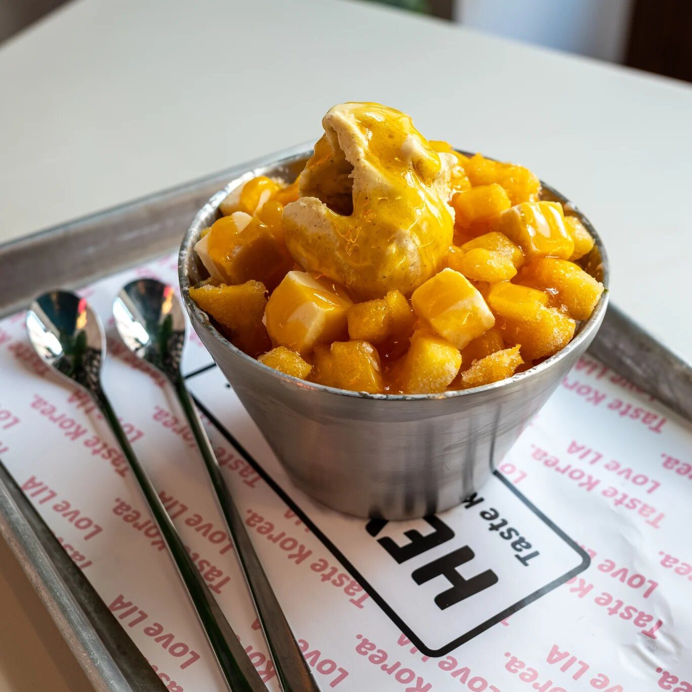 w=1367 (2) - IN München - Das Stadtmagazin Metal cup filled with cubed mango and a scoop of vanilla ice cream drizzled with mango sauce on a tray with forks nearby.