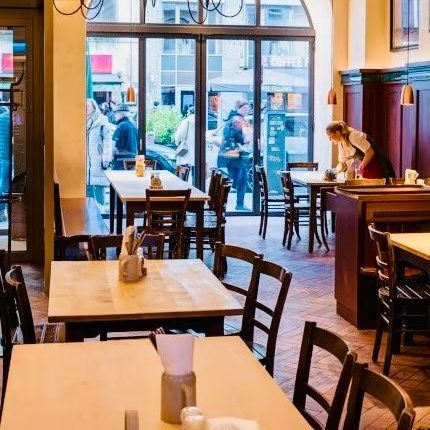 w=430 - IN München - Das Stadtmagazin Cozy cafe interior with wooden tables and chairs, a wrought-iron chandelier, and a large arched window showing people outside.