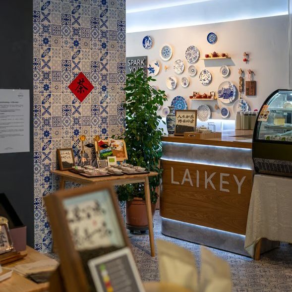 w=589 (5) - IN München - Das Stadtmagazin Cafe interior with blue-and-white tiled wall, LAIKEY counter, decorative plates on the wall, a potted plant, and pastry displays.