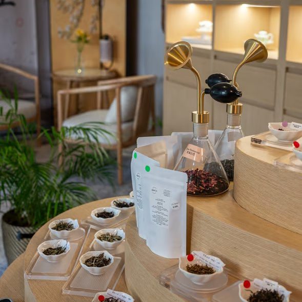 w=589 (7) - IN München - Das Stadtmagazin Tea shop counter with small bowls of loose leaf tea, two glass teapots with brass spouts, and sample bags on a curved wooden display under warm shelves of ceramic ware.
