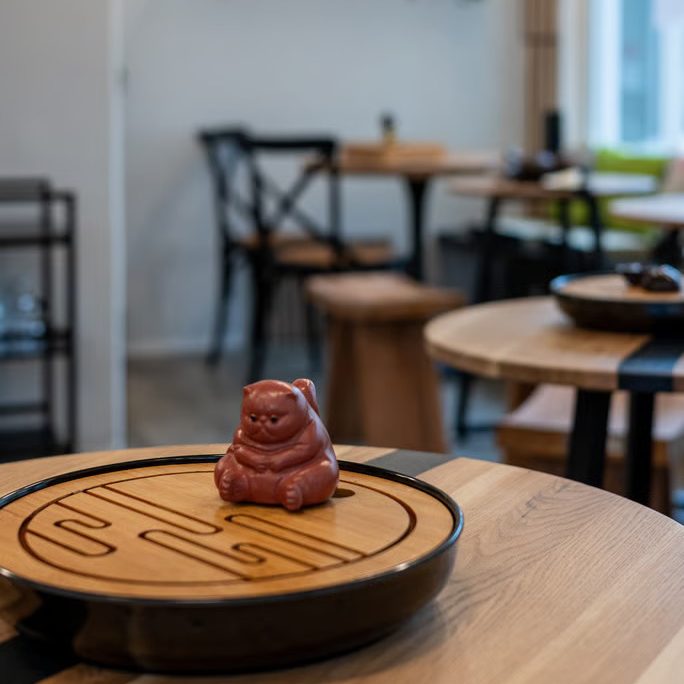 w=684 (1) - IN München - Das Stadtmagazin Chocolate Buddha statue on a round wooden platter on a cafe table, with a softly blurred dining area in the background.