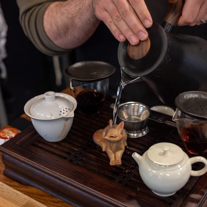w=684 (2) - IN München - Das Stadtmagazin Hands pouring hot water from a kettle onto a small wooden fox figurine on a dark tea tray, with a white teapot and cups nearby.