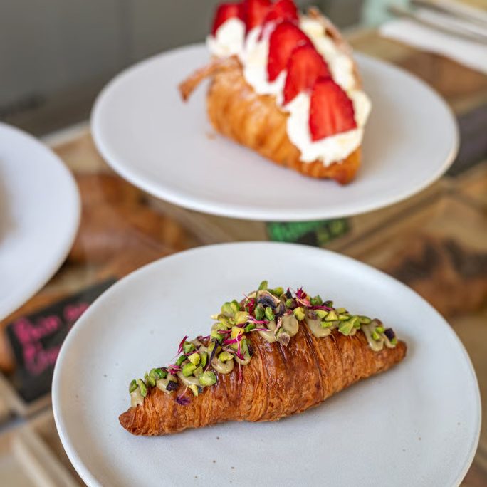 w=684 (3) - IN München - Das Stadtmagazin Croissant topped with pistachios and greens on a white plate, with a strawberry cream pastry on a separate plate in the background.