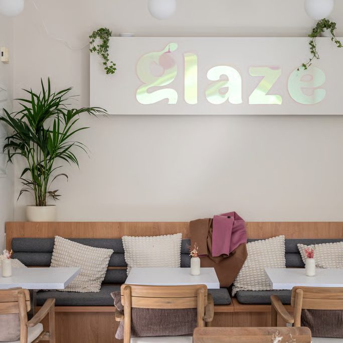 w=684 (4) - IN München - Das Stadtmagazin Cozy cafe seating area with white tables, wooden chairs, a gray cushioned bench, and a backlit 'glaze' sign on the wall.