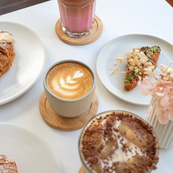 w=684 (5) - IN München - Das Stadtmagazin Brunch scene with pink layered iced drink, latte, croissants, a chocolate-topped frappé, and a vase of pink flowers on a white table.