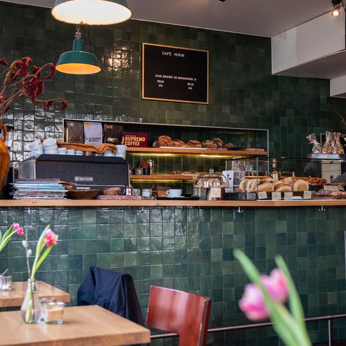 w=684 (7) - IN München - Das Stadtmagazin Cafe interior with a green tiled wall, pastry display, and chalkboard menu above the counter.