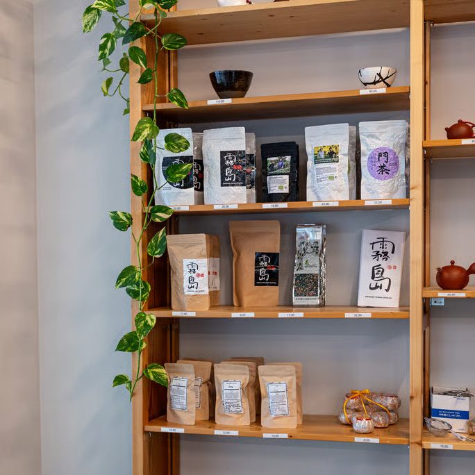 w=684 - IN München - Das Stadtmagazin Wooden shelving with a trailing plant, showcasing assorted tea bags and coffee beans in bags and tins, with price tags.
