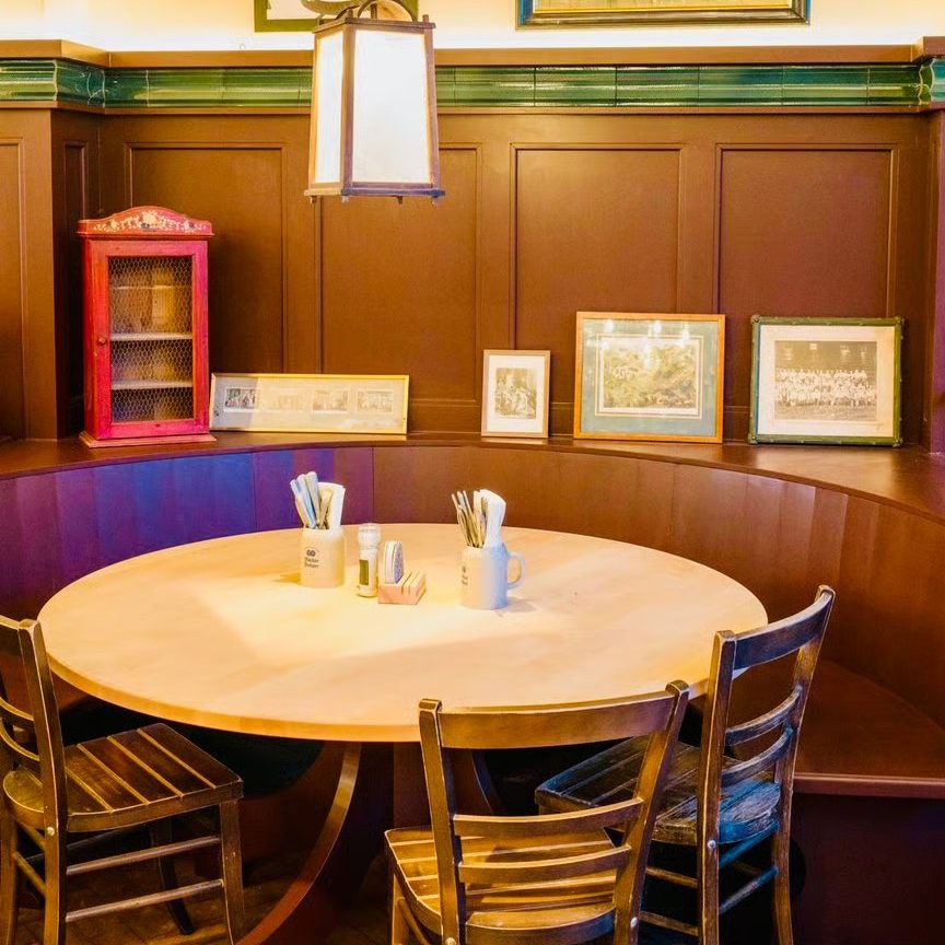 w=864 - IN München - Das Stadtmagazin Cozy dining nook with a round wooden table, four chairs, and warm pendant light in a paneled wall cafe setting, framed photos above.