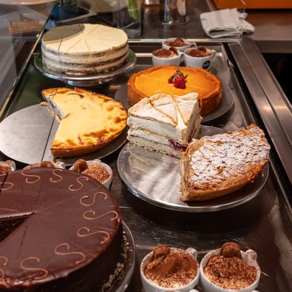 w_589-_1_ - IN München - Das Stadtmagazin Assorted cakes and pastries on display in a bakery case, including a chocolate cake, orange tart, and layered sponge cake.