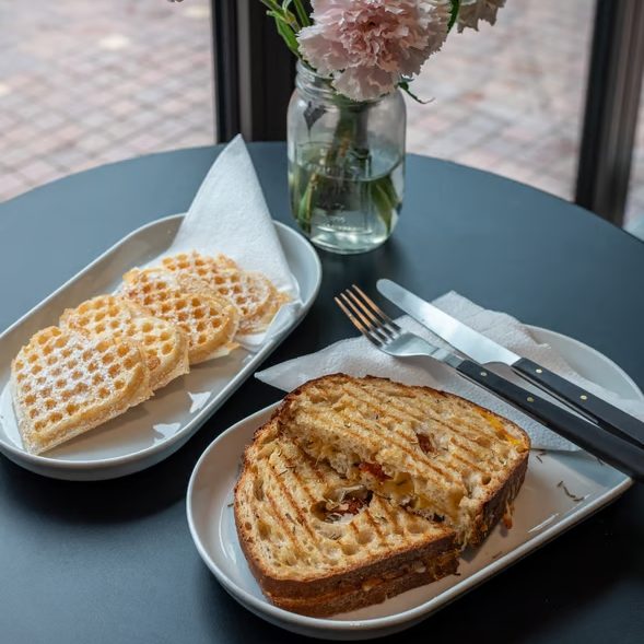w_589-_2_ - IN München - Das Stadtmagazin Table setting with a grilled sandwich on a plate, a side plate of toasted bread slices, and a vase of fresh flowers. Filetted cutlery rests on a napkin nearby.
