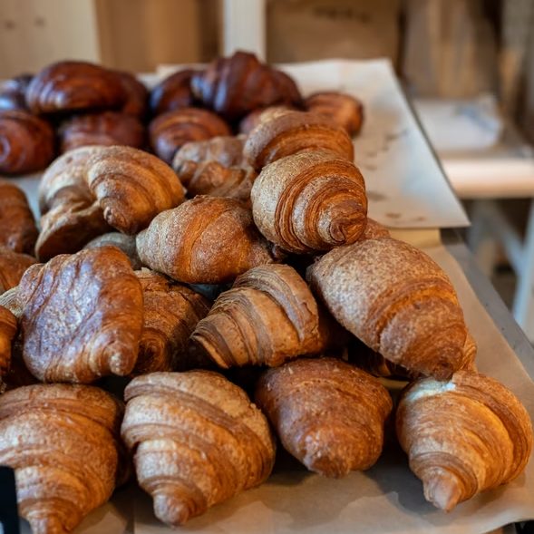 w_589-_4_ - IN München - Das Stadtmagazin Freshly baked croissants with golden flaky crust piled on parchment in a bakery display.