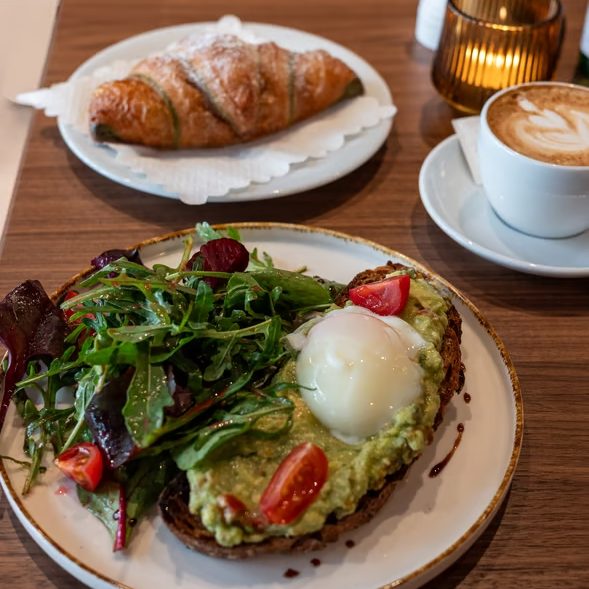 w_589 - IN München - Das Stadtmagazin Avocado toast with greens, cherry tomatoes, and a poached egg on dark bread, served with a side salad and coffee nearby.