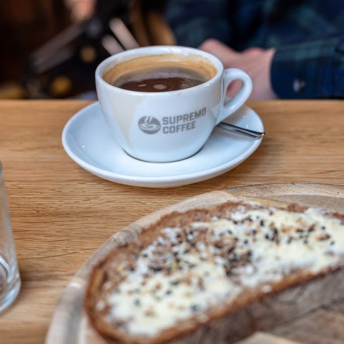 w_684-_6_ - IN München - Das Stadtmagazin White coffee cup with a Supremo Coffee logo on a saucer, set on a wooden table, with a slice of cheesy toast in the foreground.