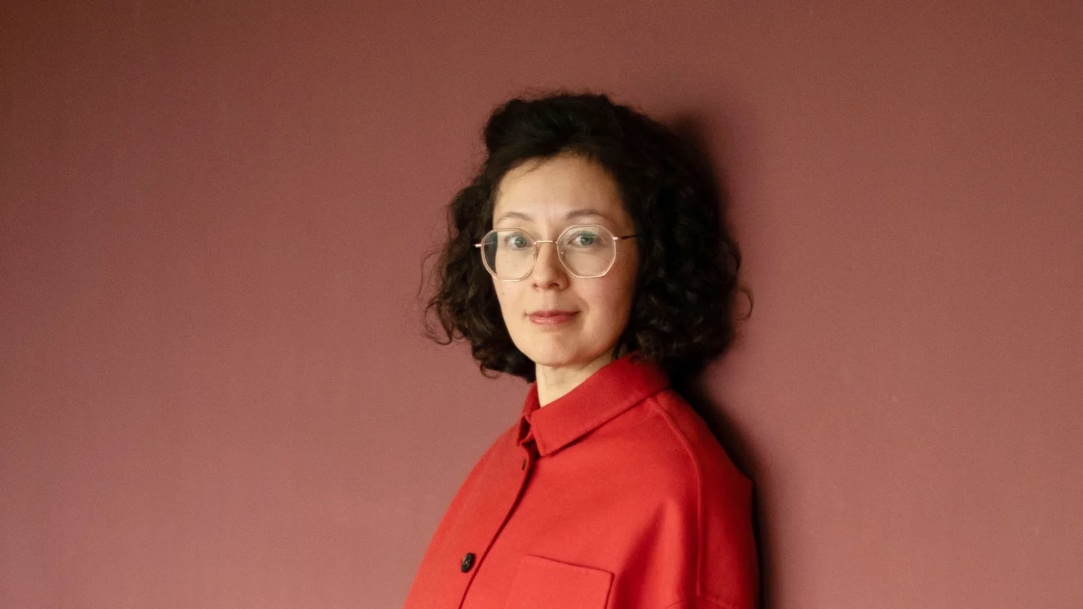 yuko kuhn - IN München - Das Stadtmagazin Woman with curly dark hair and round glasses stands against a pink wall, wearing a red button-up shirt.