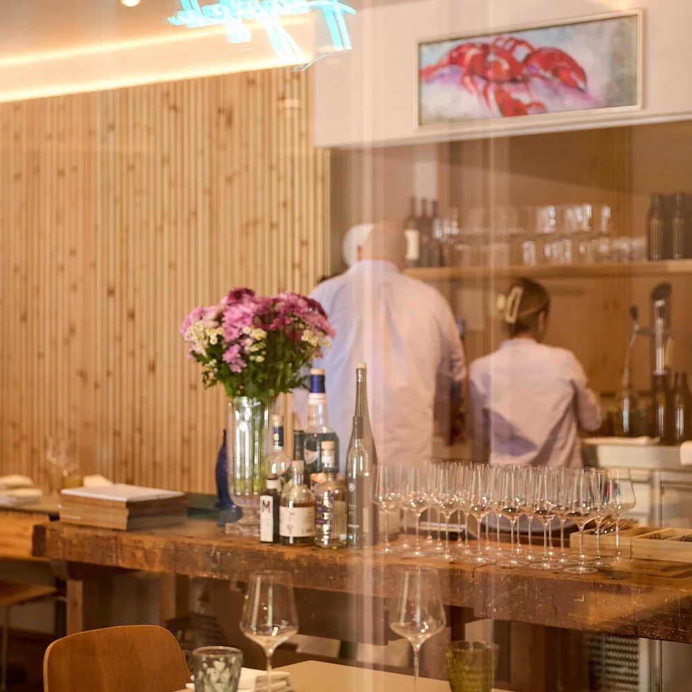 ©AnnetteSandner2 - IN München - Das Stadtmagazin Restaurant interior with a rustic wooden bar, vase of pink flowers, bottles, and rows of wine glasses ready for service in the foreground.