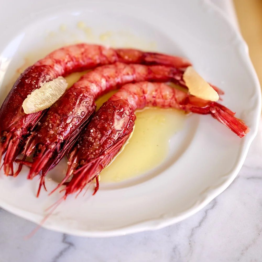 ©AnnetteSandner3 - IN München - Das Stadtmagazin Two large red shrimp tails arranged on a white plate with melted butter nearby and a lemon slice garnish