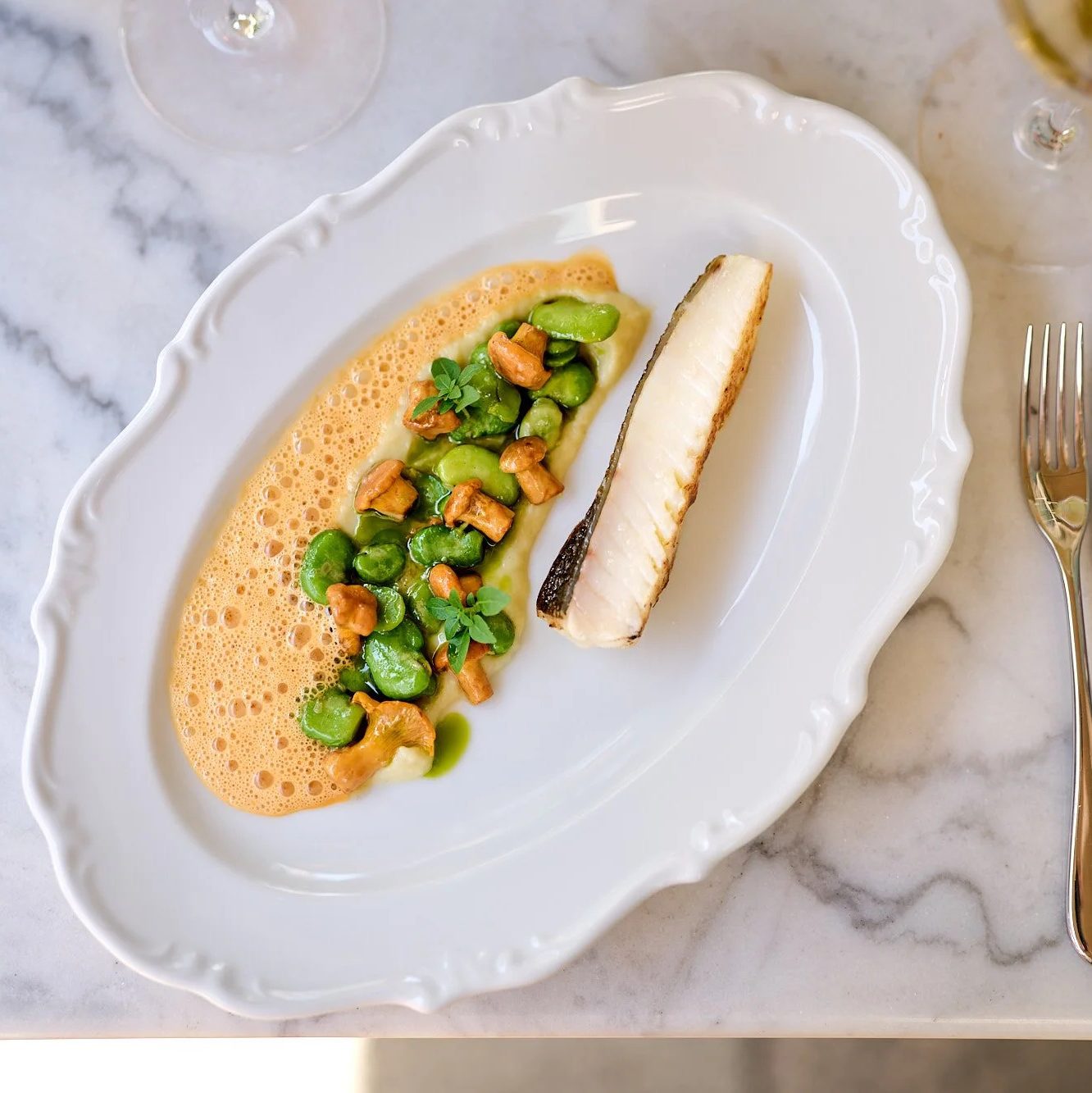 ©AnnetteSandner4 - IN München - Das Stadtmagazin Seared fish fillet on a white plate topped with green peas, almonds, and a foamy orange sauce on a marble table.