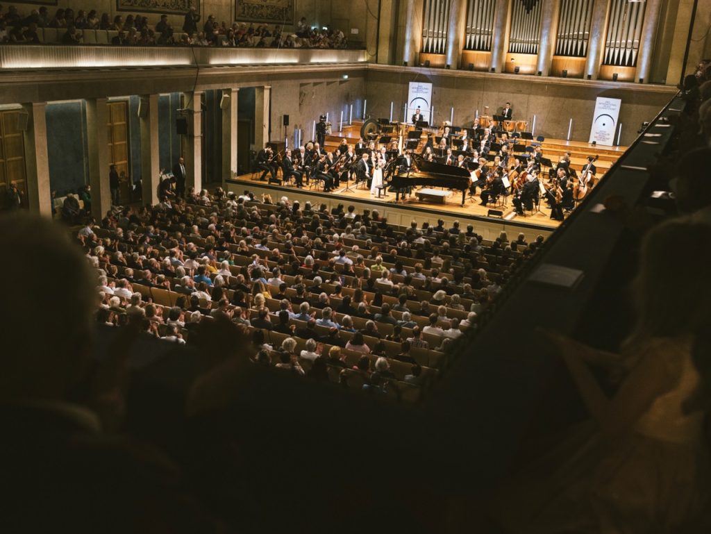 1_Herkulessaal_BRSO_IMW_ARD_2025(c)Daniel Delang_Bayerischer Rundfunk - IN München - Das Stadtmagazin Full symphony orchestra on stage in a large concert hall, with conductor and pianist, audience filling rows and balconies.