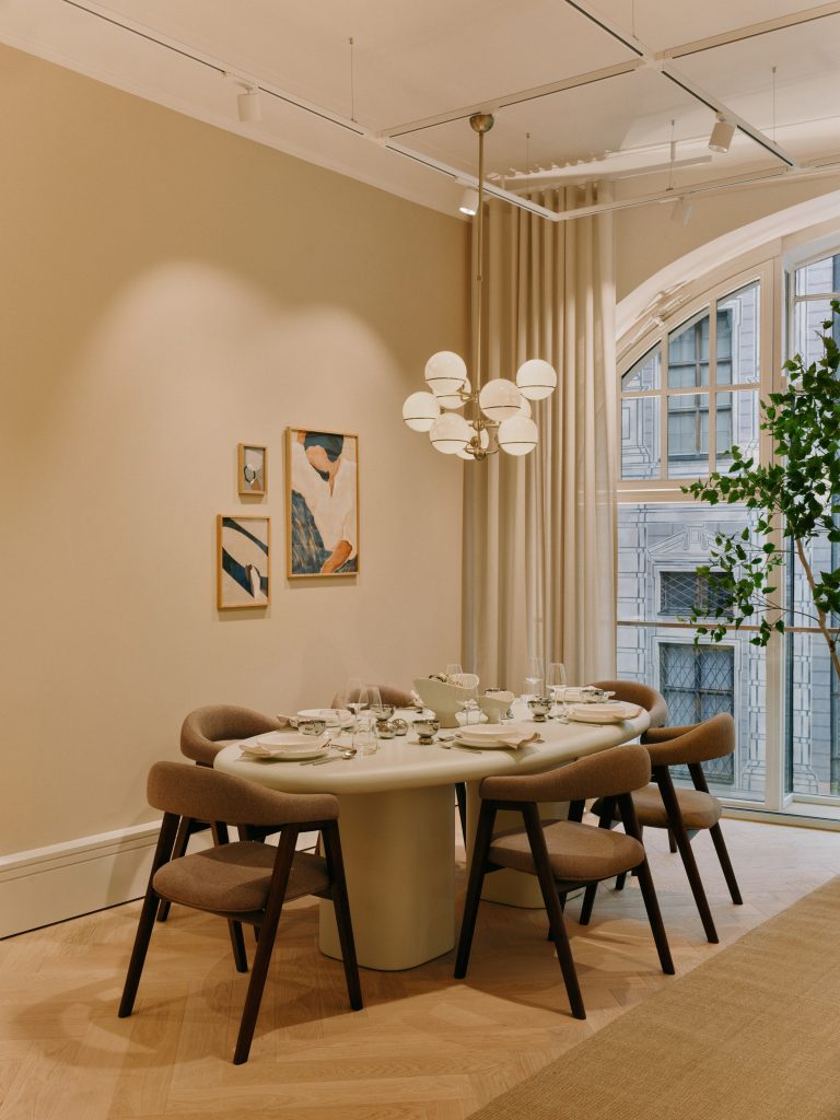 2026-07-BA-WESTWING-MUNICH-STORE-05-203 - IN München - Das Stadtmagazin Round dining table set for a meal in a softly lit room with beige walls, modern chandelier, and arched windows with light curtains.