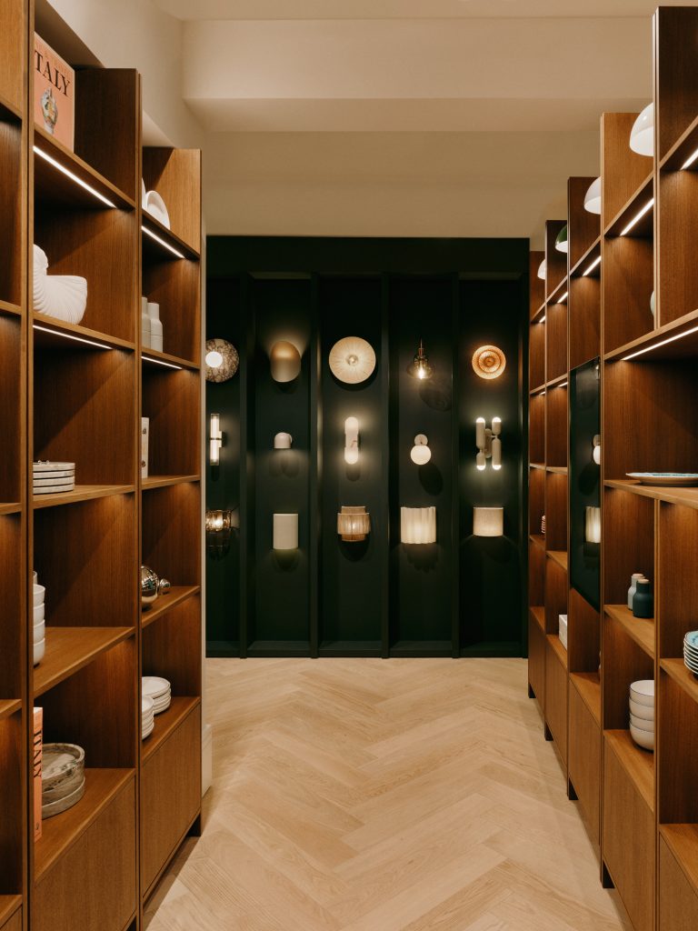2026-07-BA-WESTWING-MUNICH-STORE-08-269 - IN München - Das Stadtmagazin A showroom aisle lined with tall wooden shelves displaying plates, vases and lighting fixtures against a dark back wall.