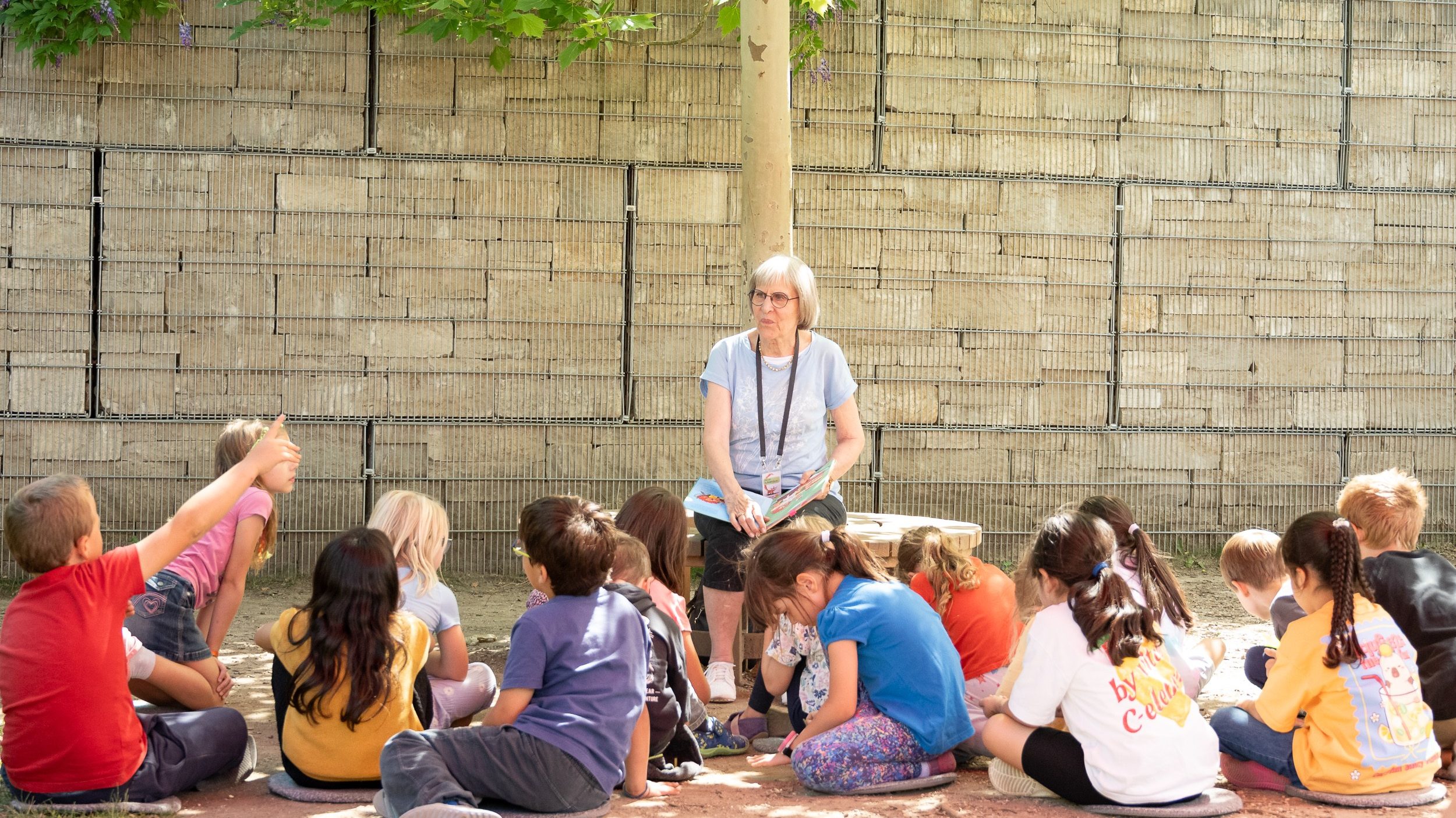 20260720_IrisKlein_Lesefchse-18 - IN München - Das Stadtmagazin Older woman with glasses reads a colorful book to a group of children seated on the ground outdoors near a stone wall.