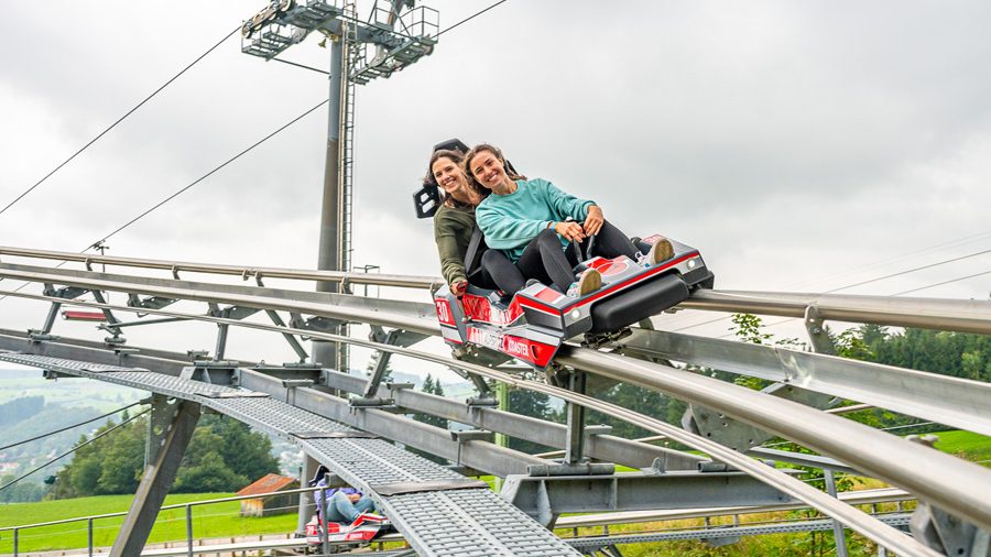 Alpspitz-COASTER - IN München - Das Stadtmagazin Two people ride a red roller coaster car along a metal track, smiling at the camera with a cloudy sky above.