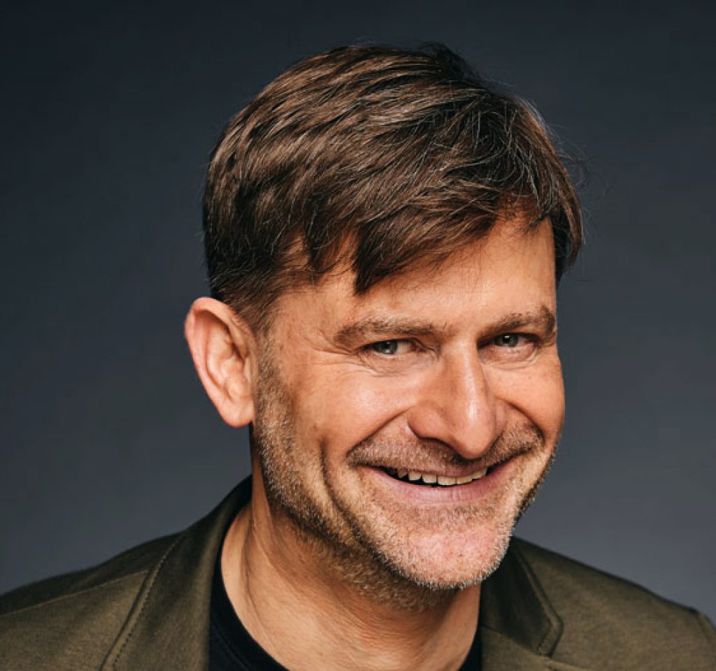 Altinger - IN München - Das Stadtmagazin Smiling man with short brown hair and light stubble, wearing an olive jacket against a dark gray background (head-and-shoulders portrait).
