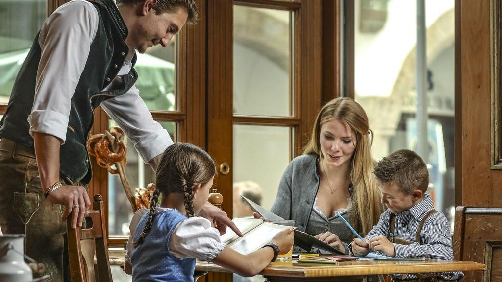 Restaurant scene: a waiter leans over a wooden table to help a mother and her two children with drawings and reading materials in a cozy cafe.