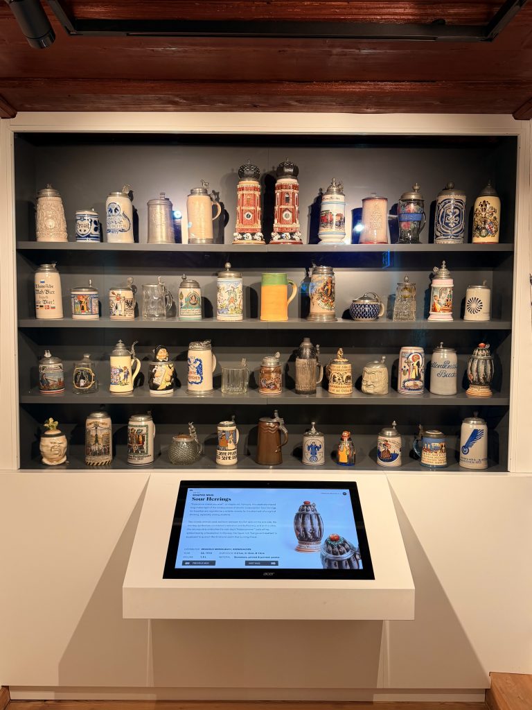 Biermuseum-muenchen-bierkruege - IN München - Das Stadtmagazin Keferloher Maßkrüge im Biermuseum München hinter einer Glaswand, davor ein Display auf dem man die einzelnen Krüge auswählen und die Geschichte nachlesen kann