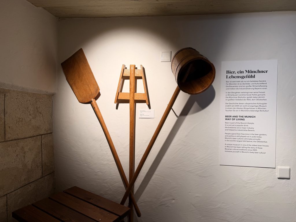 Biermuseum-muenchen-brauerwerkzeuge - IN München - Das Stadtmagazin Brauerwerkzeuge aus Holz, die ineinander verschränkt an einer weißen Wand befestigt und beleuchtet sind im Biermuseum München