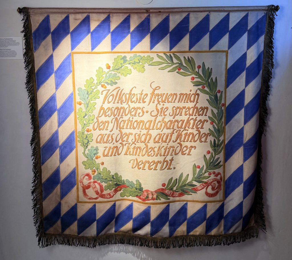 Biermuseum-muenchen-oktoberfest-fahne - IN München - Das Stadtmagazin Banner zum Oktoberfest, außen blau weiß und mit goldener Schrift im Zentrum