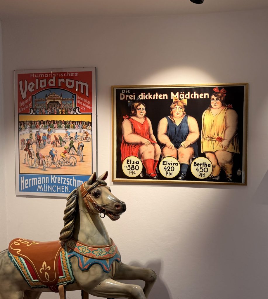 Biermuseum-muenchen-oktoberfest-schausteller-reklame - IN München - Das Stadtmagazin historische Schaustellerreklamen an einer weißen Wand sowie Exponate wie ein Clown oder Karussellpferd