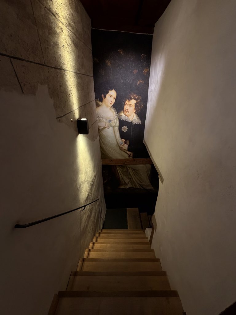 Biermuseum-muenchen-treppenhaus-zwischen-ausstellungsräumen - IN München - Das Stadtmagazin Holztreppe im Biermuseum München die nach unten führt, auf der hohen, schmalen Wand darüber erstreckt sich eine altes Gemälde, dass eine Frau und einen mann zeigt