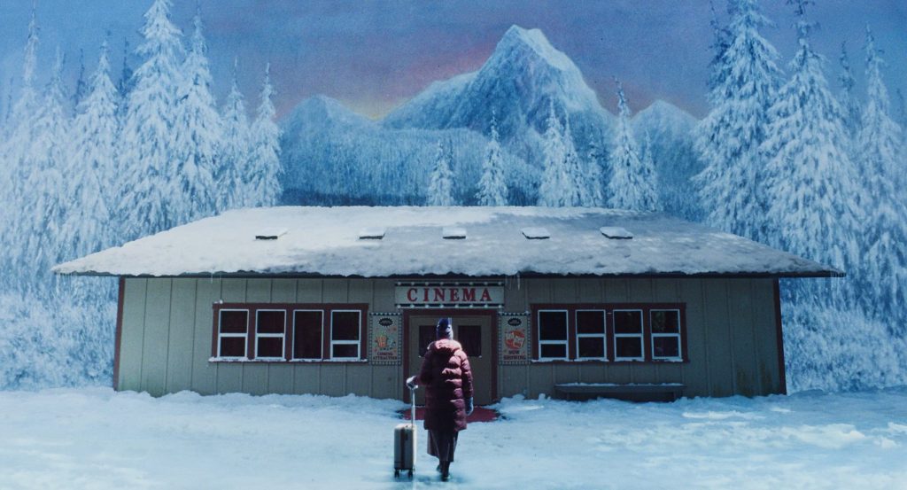 Teenager sex and death - IN München - Das Stadtmagazin Snow-covered cinema in a pine forest; a person in a red coat pulls a suitcase toward the entrance.