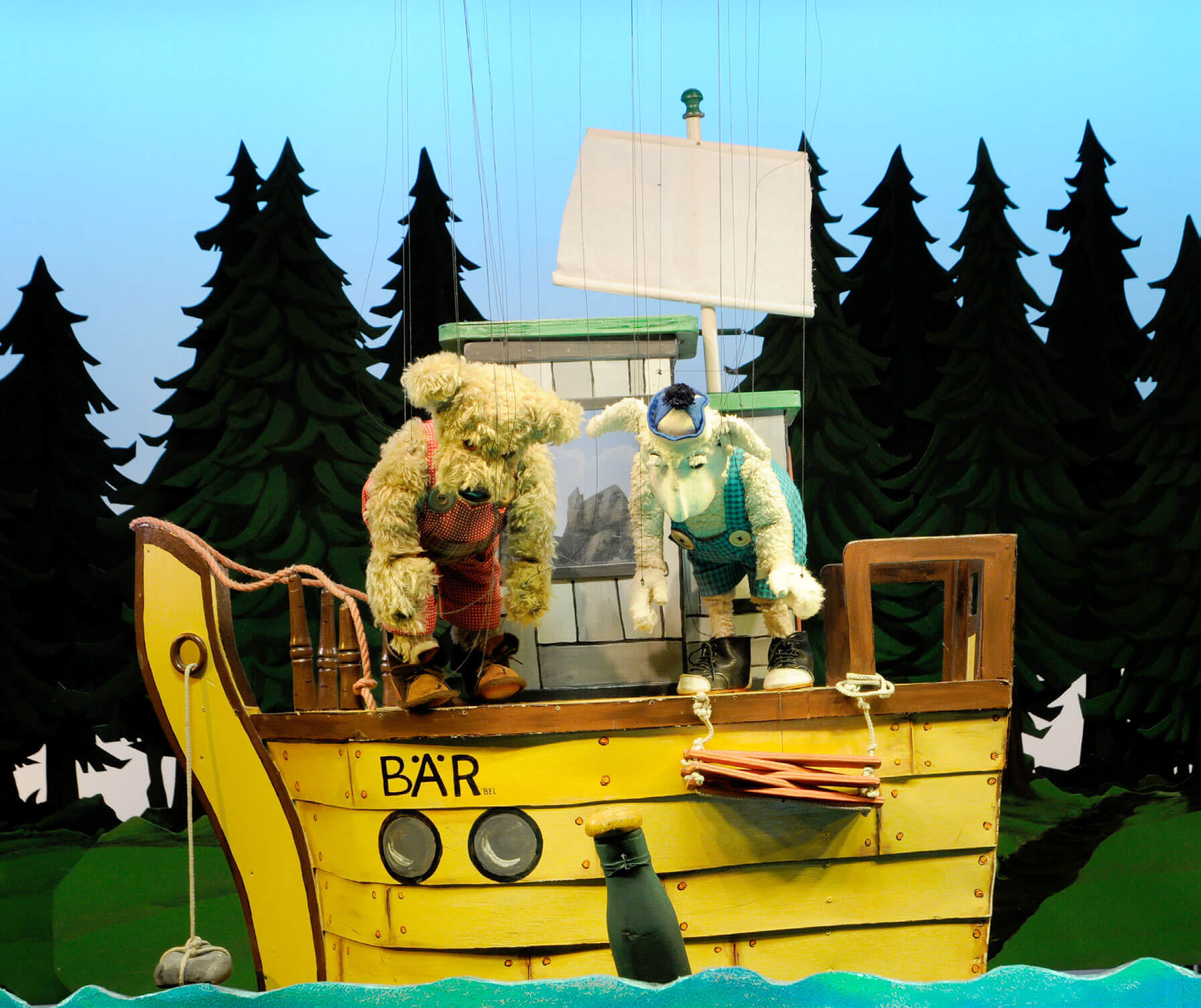 Two furry puppets stand on a bright yellow pirate-style ship stage, with ropes, a green deck, and a forest silhouette backdrop behind them.