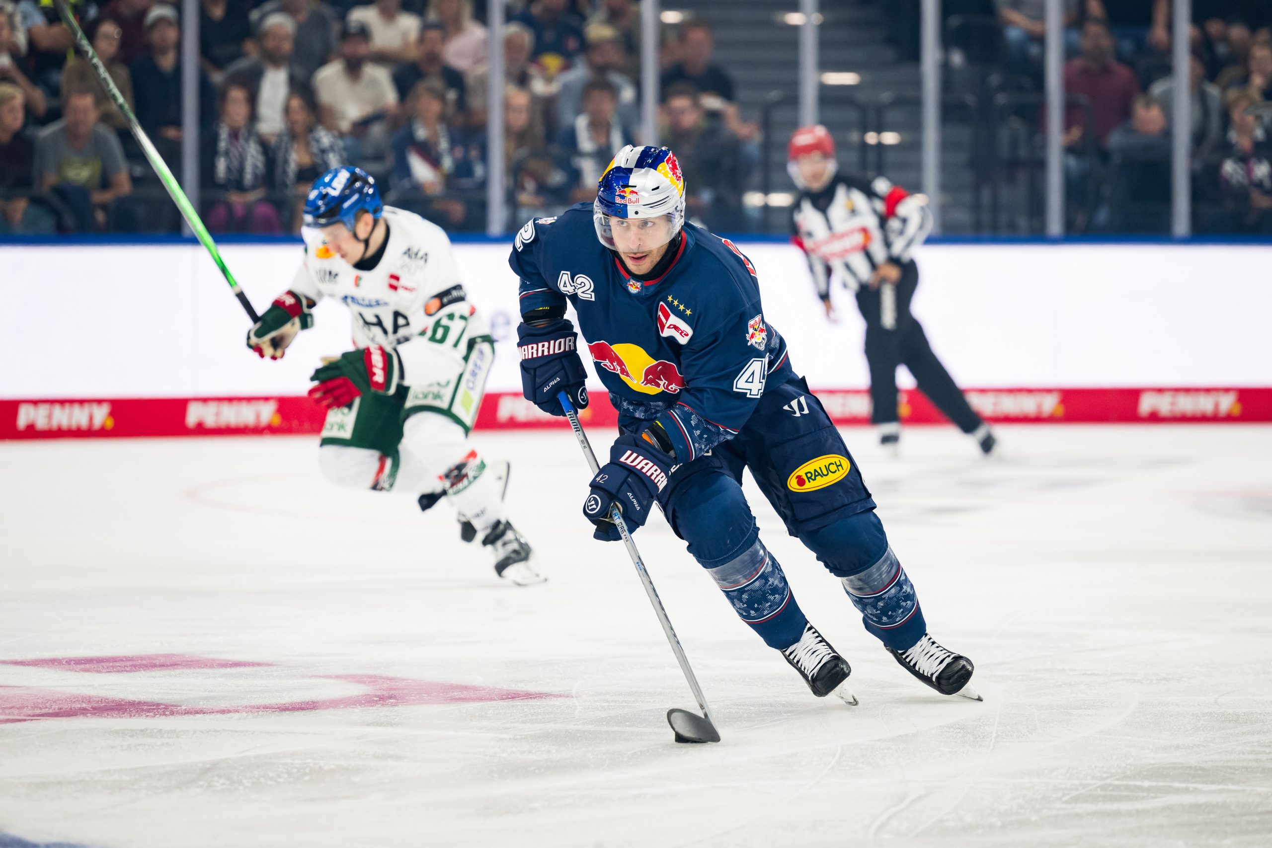 Ice hockey player in a blue jersey with Red Bull logo (#42) skating forward with the puck, while a white-jersey opponent and fans watch from the stands behind.