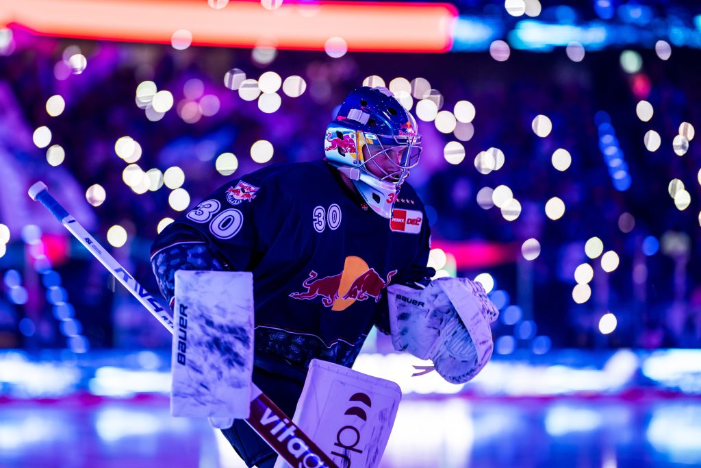 Red Bull Munchen gegen Straubing Tigers - PENNY DEL - IN München - Das Stadtmagazin Ice hockey goalie in a dark jersey with Red Bull branding, ready to block a shot as colorful arena lights glow in the background.
