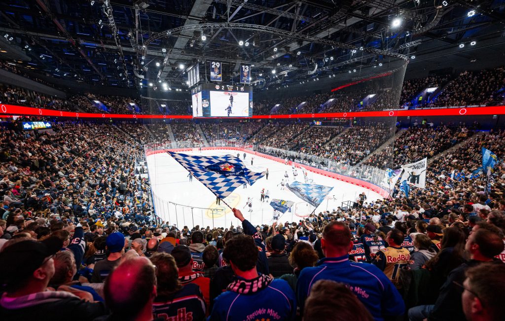 EHC Red Bull Munchen gegen Adler Mannheim - PENNY DEL Halbfinale Spiel 2 - IN München - Das Stadtmagazin Packed hockey arena with fans waving blue-and-white flags over the ice rink and a large LED scoreboard overhead.