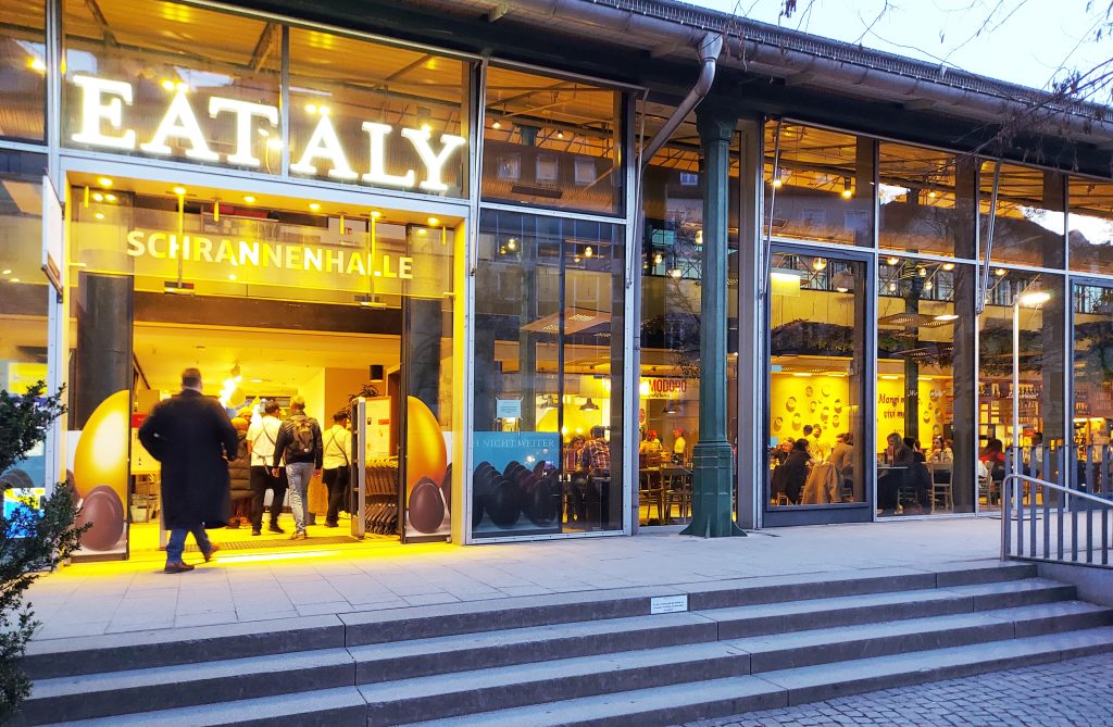 Eataly_Schrannenhalle_Muenchen - IN München - Das Stadtmagazin Eataly storefront with a large neon sign and customers inside seen through glass façade at dusk